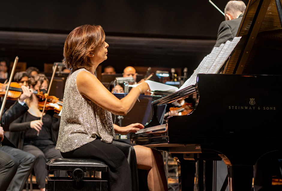 A white women with short, brown hair, wearing a silver top and black skirt, playing a Steinway piano in front of an orchestra, with violinists playing in the background.